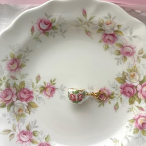 May include: A small, decorative teacup charm with a gold clasp rests on a white plate. The plate is adorned with a floral pattern of pink and white roses and green leaves. A pink ribbon is in the background.
