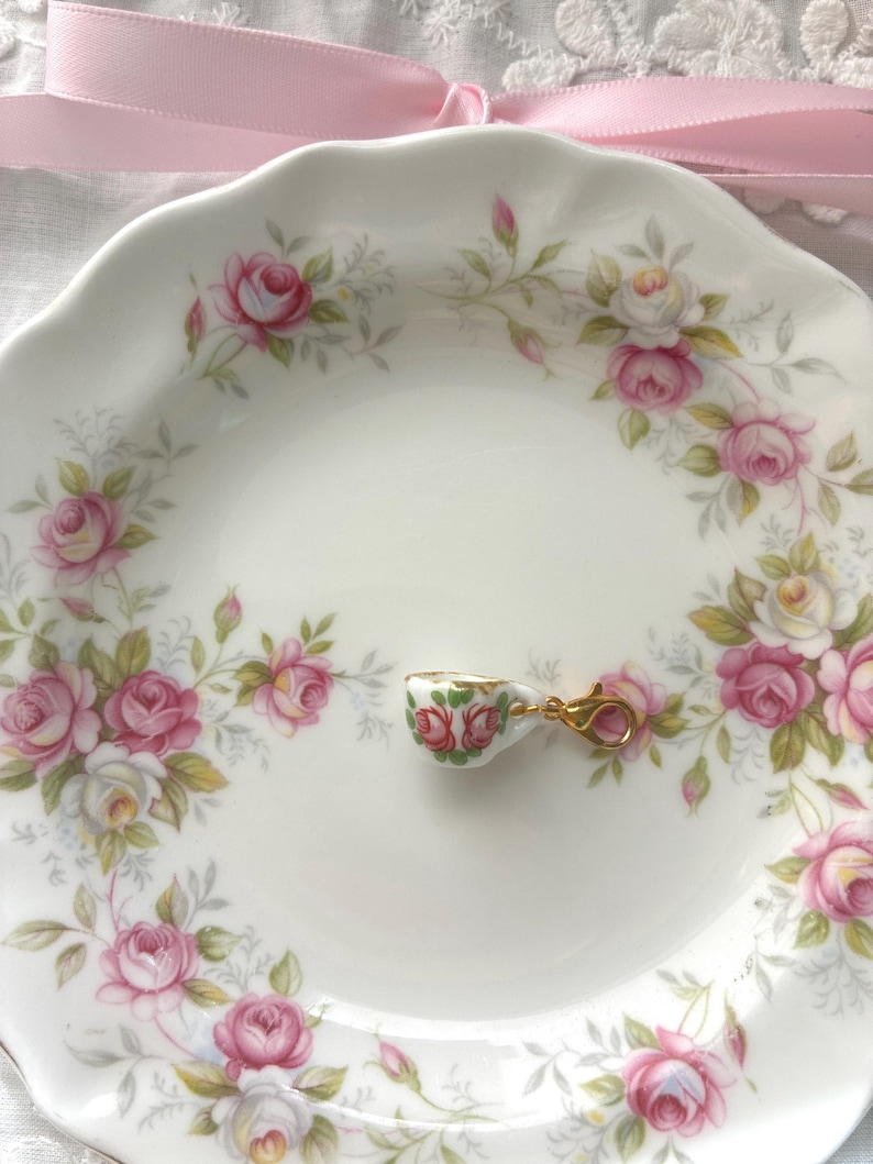 May include: A small, decorative teacup charm with a gold clasp rests on a white plate. The plate is adorned with a floral pattern of pink and white roses and green leaves. A pink ribbon is in the background.