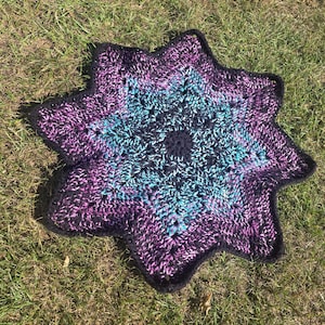 May include: A star-shaped crochet mat featuring a variegated yarn pattern in shades of deep purple, bright turquoise, and black. The center is a solid black circle, with the star points radiating outward in a textured, multi-colored stitch pattern.
