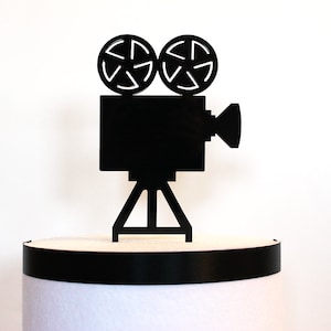 May include: Black silhouette of a vintage movie camera with two spools on top and a tripod stand. The camera is on a black and white cake stand.
