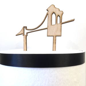 Puede incluir: Un adorno para pastel de madera con forma de Golden Gate Bridge. El adorno es de color marr&oacute;n y tiene una cinta negra alrededor de la base.