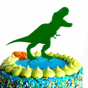 May include: A blue and green frosted cake decorated with green and orange sprinkles and a green dinosaur cake topper.