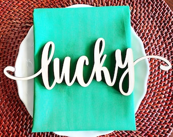 May include: A white wooden sign with the word "lucky" written in cursive script. The sign is placed on a green napkin on a white plate. The plate is on top of a brown wicker placemat.