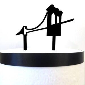 Puede incluir: Topper de pastel en forma de puente negro. El puente tiene una torre a cada lado y un cable que conecta las torres. El topper de pastel est&aacute; sobre un pastel blanco con una cinta negra.