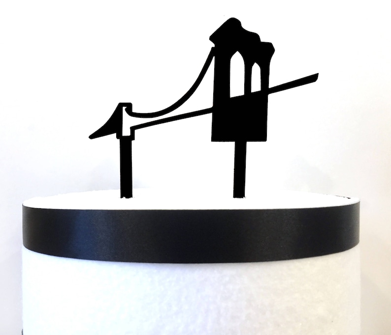 Puede incluir: Topper de pastel en forma de puente negro. El puente tiene una torre a cada lado y un cable que conecta las torres. El topper de pastel est&aacute; sobre un pastel blanco con una cinta negra.