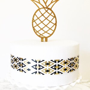 May include: A white cake decorated with a gold glitter pineapple cake topper and a black and gold geometric patterned ribbon. The cake is on a white cake stand.