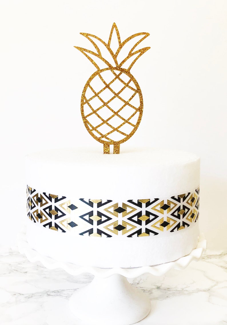 May include: A white cake decorated with a gold glitter pineapple cake topper and a black and gold geometric patterned ribbon. The cake is on a white cake stand.