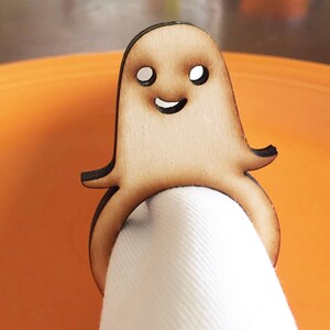 May include: A wooden ghost napkin ring with a smiling face. The ring is brown and has a white napkin inside.