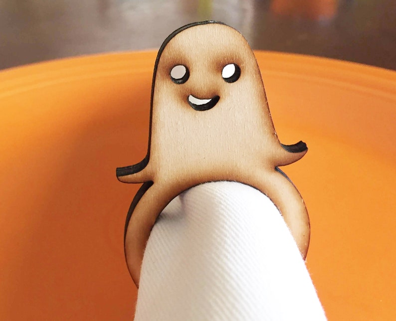 May include: A wooden ghost napkin ring with a smiling face. The ring is brown and has a white napkin inside.