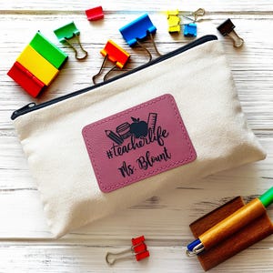 Op de afbeelding: Een beige canvas etui met een zwarte rits en een roze leren patch. De patch heeft de tekst "#teacherlife Ms. Blount" en afbeeldingen met een leraren thema. Gekleurde paperclips en een pen zijn ook zichtbaar.