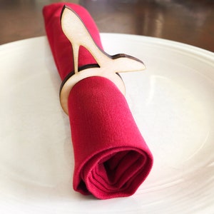 May include: A wooden high heel shoe napkin ring. The ring is made of light brown wood and is shaped like a high heel shoe. The ring is holding a red napkin.