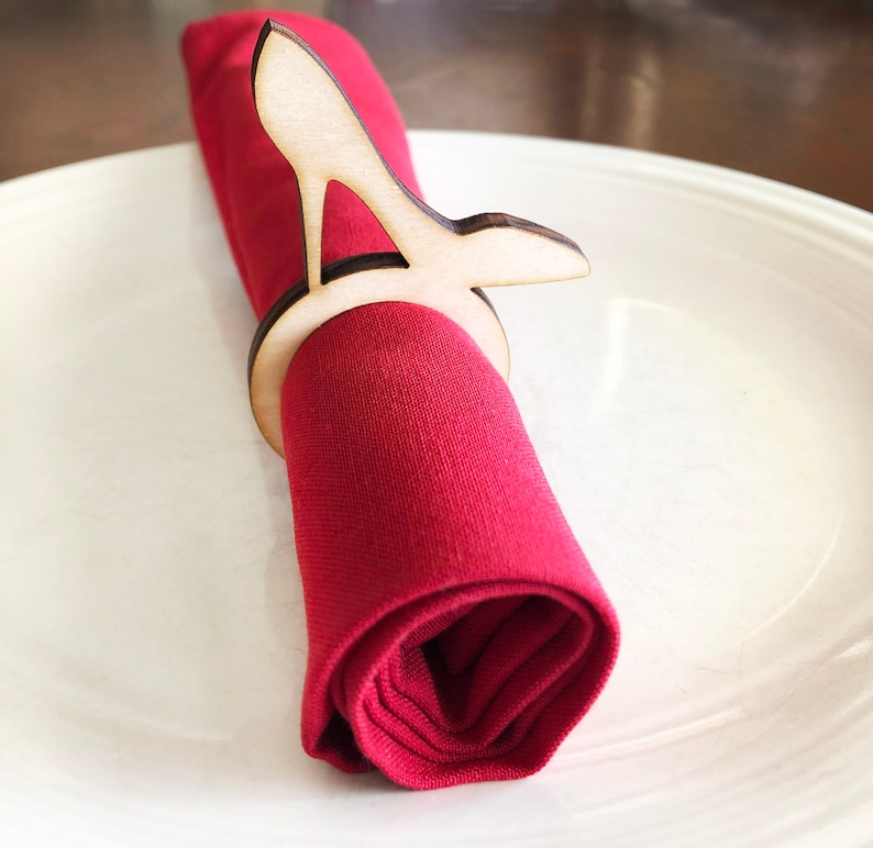 May include: A wooden high heel shoe napkin ring. The ring is made of light brown wood and is shaped like a high heel shoe. The ring is holding a red napkin.