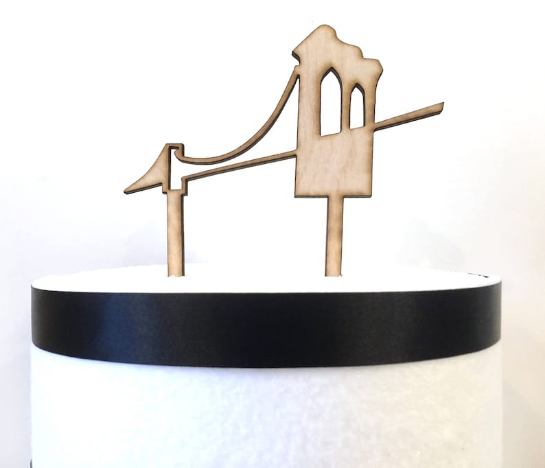 Puede incluir: Un adorno para pastel de madera con forma de Golden Gate Bridge. El adorno es de color marr&oacute;n y tiene una cinta negra alrededor de la base.