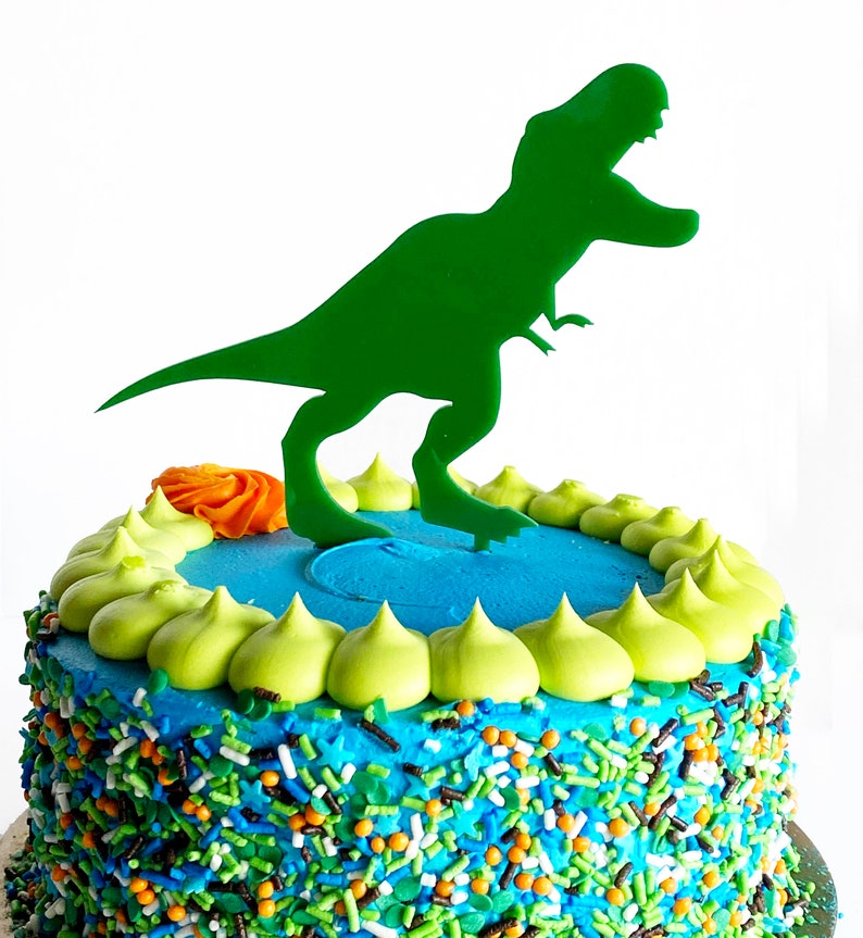 May include: A blue and green frosted cake decorated with green and orange sprinkles and a green dinosaur cake topper.