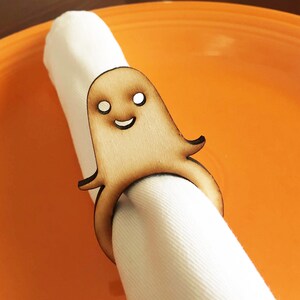 May include: A wooden ghost napkin ring with a smiling face. The ghost is light brown and has white eyes. The napkin ring is on a white napkin.
