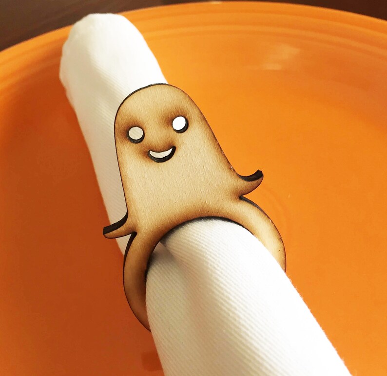 May include: A wooden ghost napkin ring with a smiling face. The ghost is light brown and has white eyes. The napkin ring is on a white napkin.