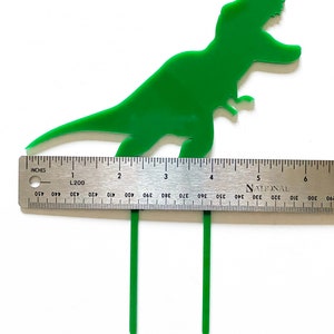 May include: Green plastic dinosaur cake topper with two green plastic sticks. The dinosaur is approximately 4 inches tall. The sticks are approximately 4 inches long.