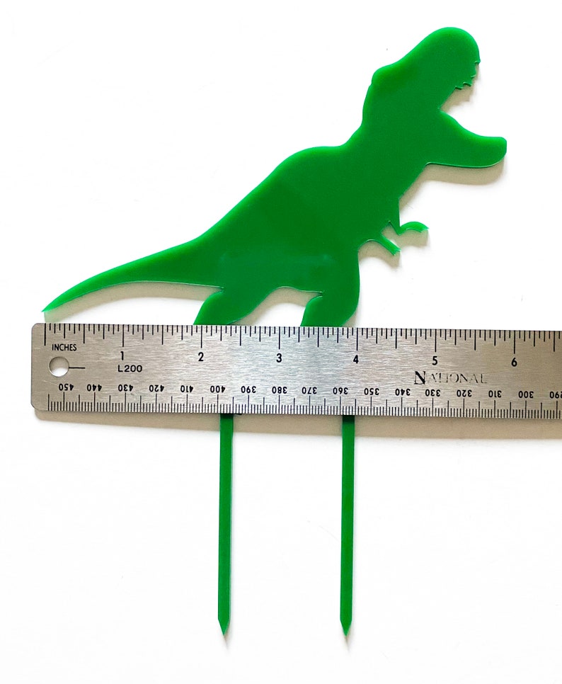 May include: Green plastic dinosaur cake topper with two green plastic sticks. The dinosaur is approximately 4 inches tall. The sticks are approximately 4 inches long.