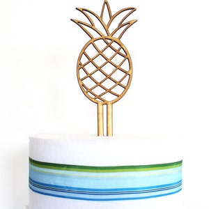 May include: A wooden pineapple cake topper with a brown color. The topper is on a white cake with blue and green striped ribbon.