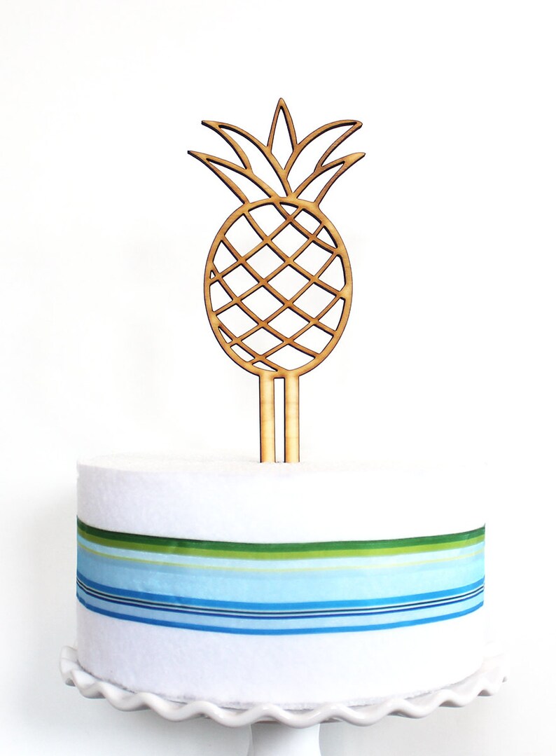 May include: A wooden pineapple cake topper with a brown color. The topper is on a white cake with blue and green striped ribbon.