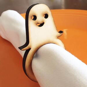 May include: Wooden ghost napkin ring with a smiling face. The ghost is brown and has a white napkin inside.
