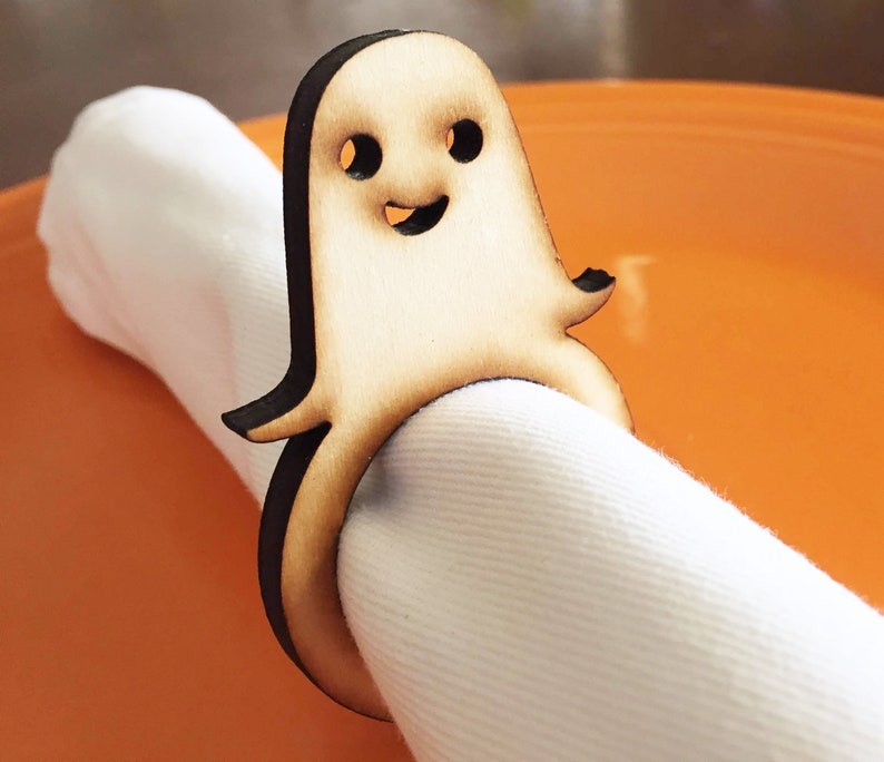 May include: Wooden ghost napkin ring with a smiling face. The ghost is brown and has a white napkin inside.