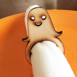 May include: Wooden ghost napkin ring with a smiling face. The ring is holding a white napkin.