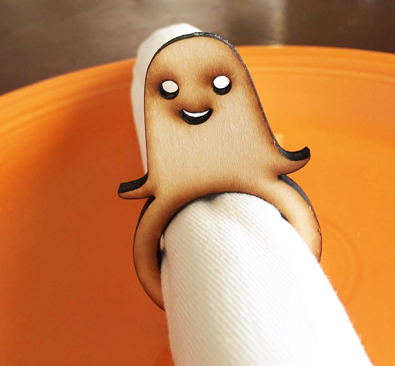 May include: Wooden ghost napkin ring with a smiling face. The ring is holding a white napkin.