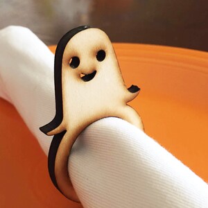 May include: A wooden ghost napkin ring with a smiling face. The ring is brown with black details and is holding a white napkin.