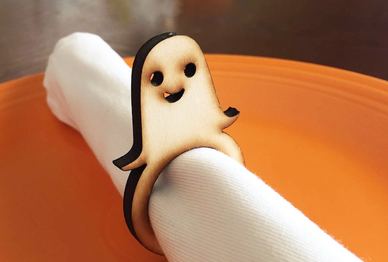 May include: A wooden ghost napkin ring with a smiling face. The ring is brown with black details and is holding a white napkin.
