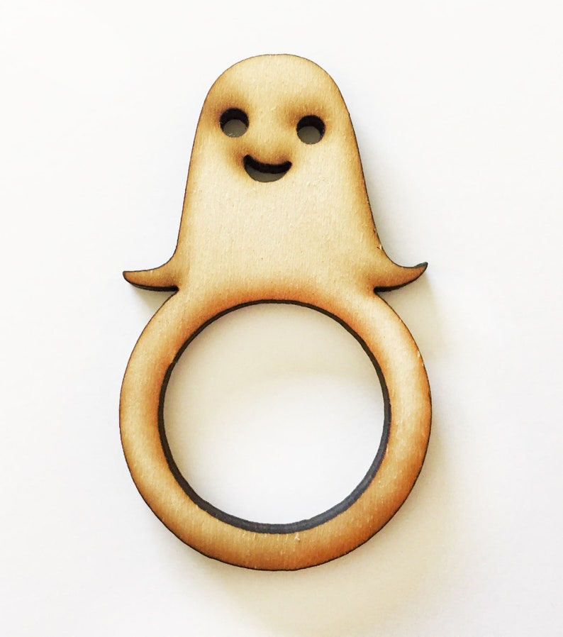 May include: A wooden ghost napkin ring with a smiling face. The ring is light brown and has a smooth finish.