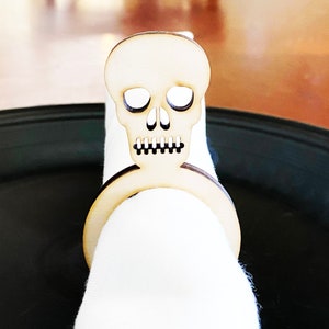 May include: A wooden skull napkin ring. The skull is carved with eye sockets, a nose, and a mouth. The ring is designed to hold a napkin in place.