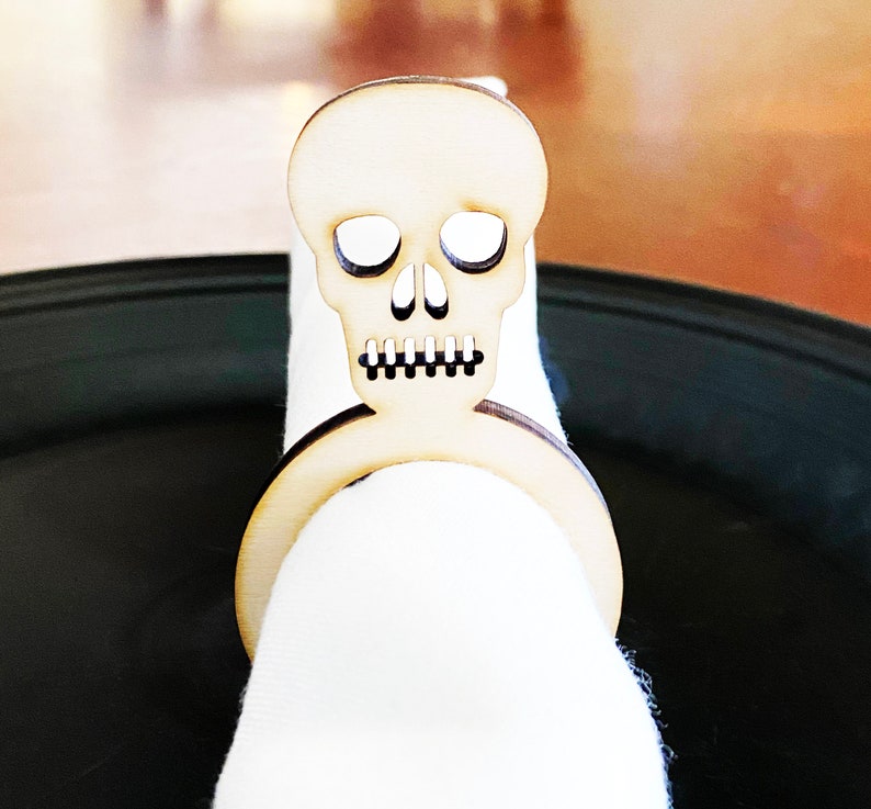 May include: A wooden skull napkin ring. The skull is carved with eye sockets, a nose, and a mouth. The ring is designed to hold a napkin in place.