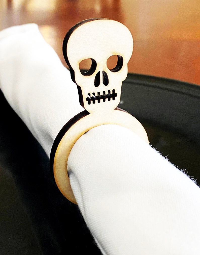 May include: Wooden skull napkin ring with a black and white skull design. The ring is made of natural wood and has a smooth finish.