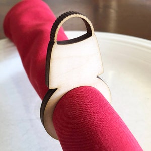 以下が含まれることがあります： ハンドバッグの形をした木製ナプキンリング。リングは薄い木でできており、茶色の染色が施され、切り抜かれたハンドルが付いています。リングは赤いナプキンを持っています。