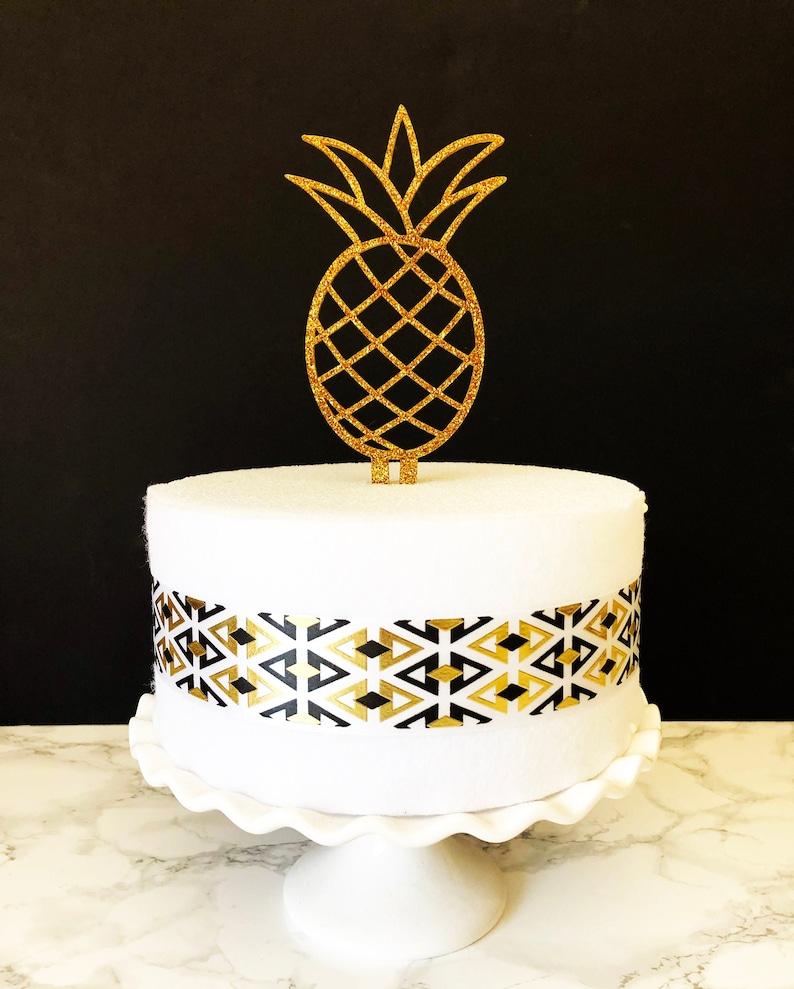 May include: A white cake with a black and gold geometric pattern around the bottom tier. A gold glitter pineapple cake topper sits on top of the cake.