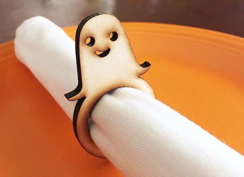 May include: A wooden ghost napkin ring with a smiling face. The ring is holding a white napkin. The ring is on a bright orange surface.