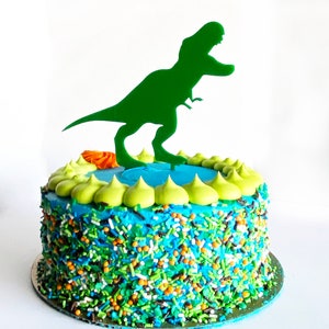 May include: A blue and green frosted cake decorated with colorful sprinkles and a green dinosaur silhouette topper.