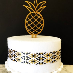 May include: A white cake with a black and gold geometric patterned ribbon around the bottom tier. A gold glitter pineapple cake topper sits on top of the cake.
