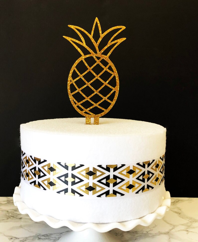 May include: A white cake with a black and gold geometric patterned ribbon around the bottom tier. A gold glitter pineapple cake topper sits on top of the cake.