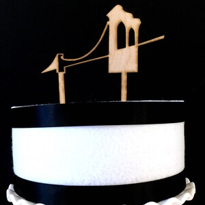 Puede incluir: Un adorno para pastel de madera con forma de Golden Gate Bridge. El adorno es marr&oacute;n y tiene un fondo blanco.