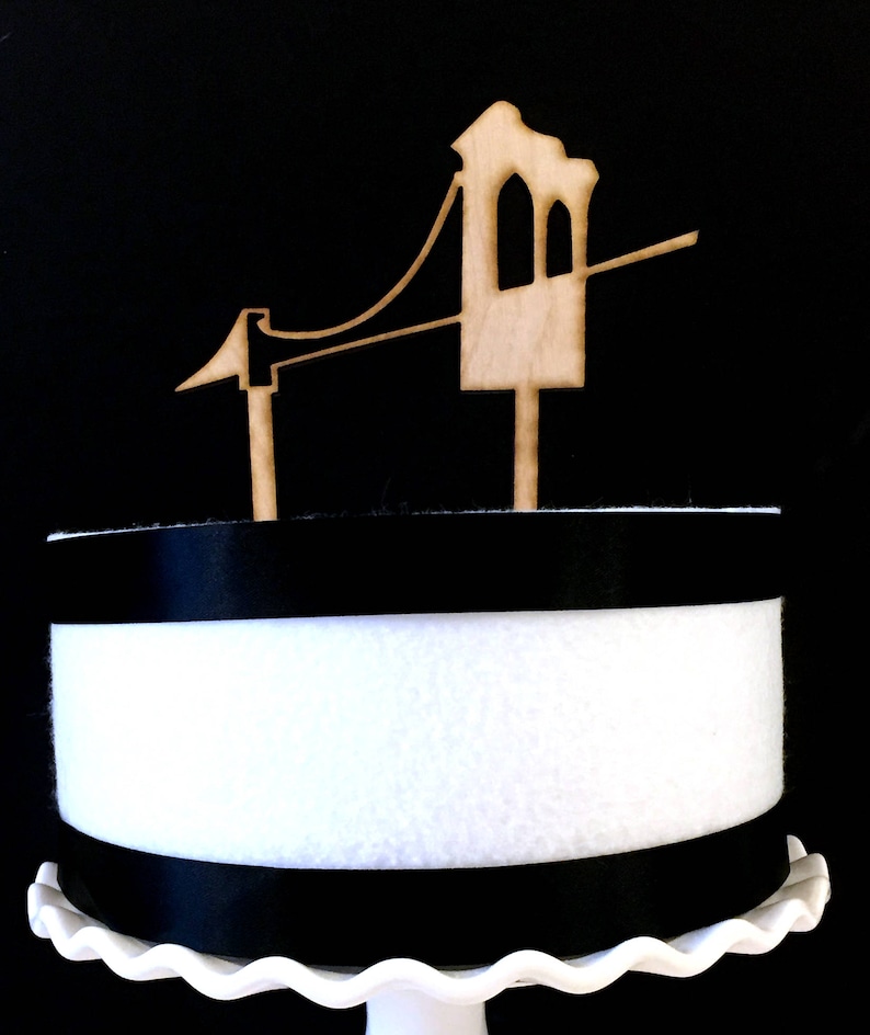 Puede incluir: Un adorno para pastel de madera con forma de Golden Gate Bridge. El adorno es marr&oacute;n y tiene un fondo blanco.