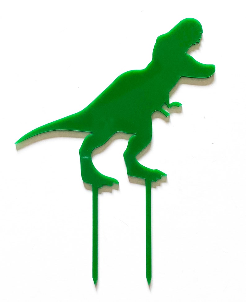 May include: Green dinosaur cake topper with two green sticks. The dinosaur is standing on its hind legs with its mouth open.