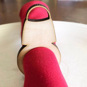 以下が含まれることがあります： 茶色の仕上げをしたハンドバッグの形をした木製ナプキンリング。リングは赤いナプキンを持っています。