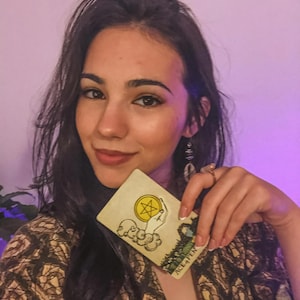 May include: A tarot card with the text "ACE OF PENTACLES" is held up. The card features a hand holding a pentacle, with a landscape scene in the background. The card has a yellow and green color scheme.