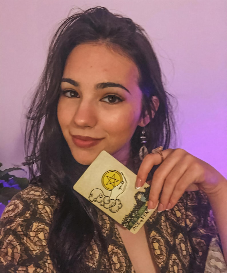 May include: A tarot card with the text "ACE OF PENTACLES" is held up. The card features a hand holding a pentacle, with a landscape scene in the background. The card has a yellow and green color scheme.