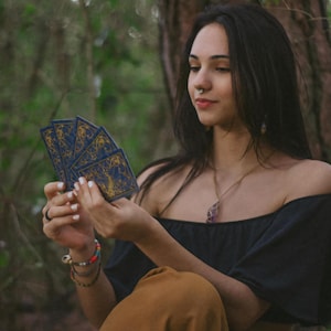 May include: A person holding a fan of tarot cards with a blue and gold design. The person is wearing a black off-the-shoulder top and brown pants. The background is a forest setting.