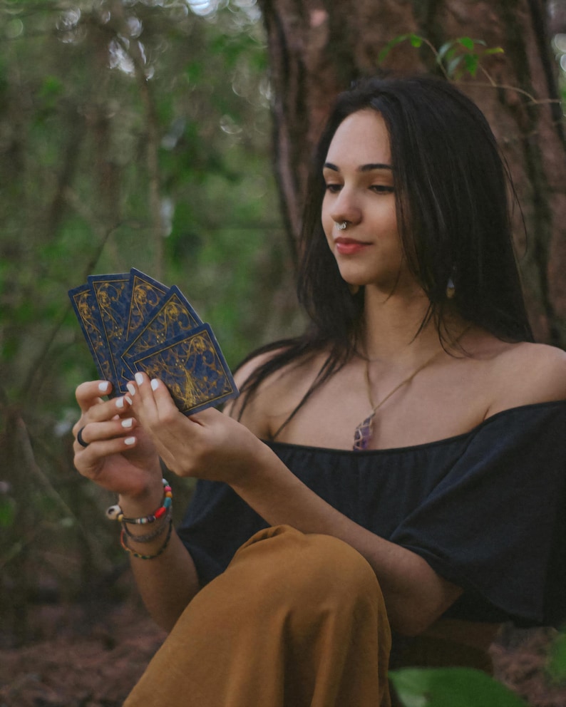 May include: A person holding a fan of tarot cards with a blue and gold design. The person is wearing a black off-the-shoulder top and brown pants. The background is a forest setting.