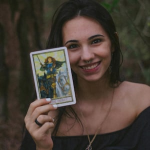 May include: A woman holding a tarot card depicting a warrior on a chariot pulled by two horses. The card has a white border and the text "THE CHARIOT" at the bottom. The woman is smiling and wearing a black top and a necklace.