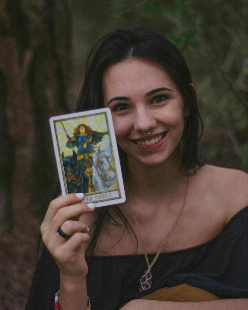 May include: A woman holding a tarot card depicting a warrior on a chariot pulled by two horses. The card has a white border and the text "THE CHARIOT" at the bottom. The woman is smiling and wearing a black top and a necklace.